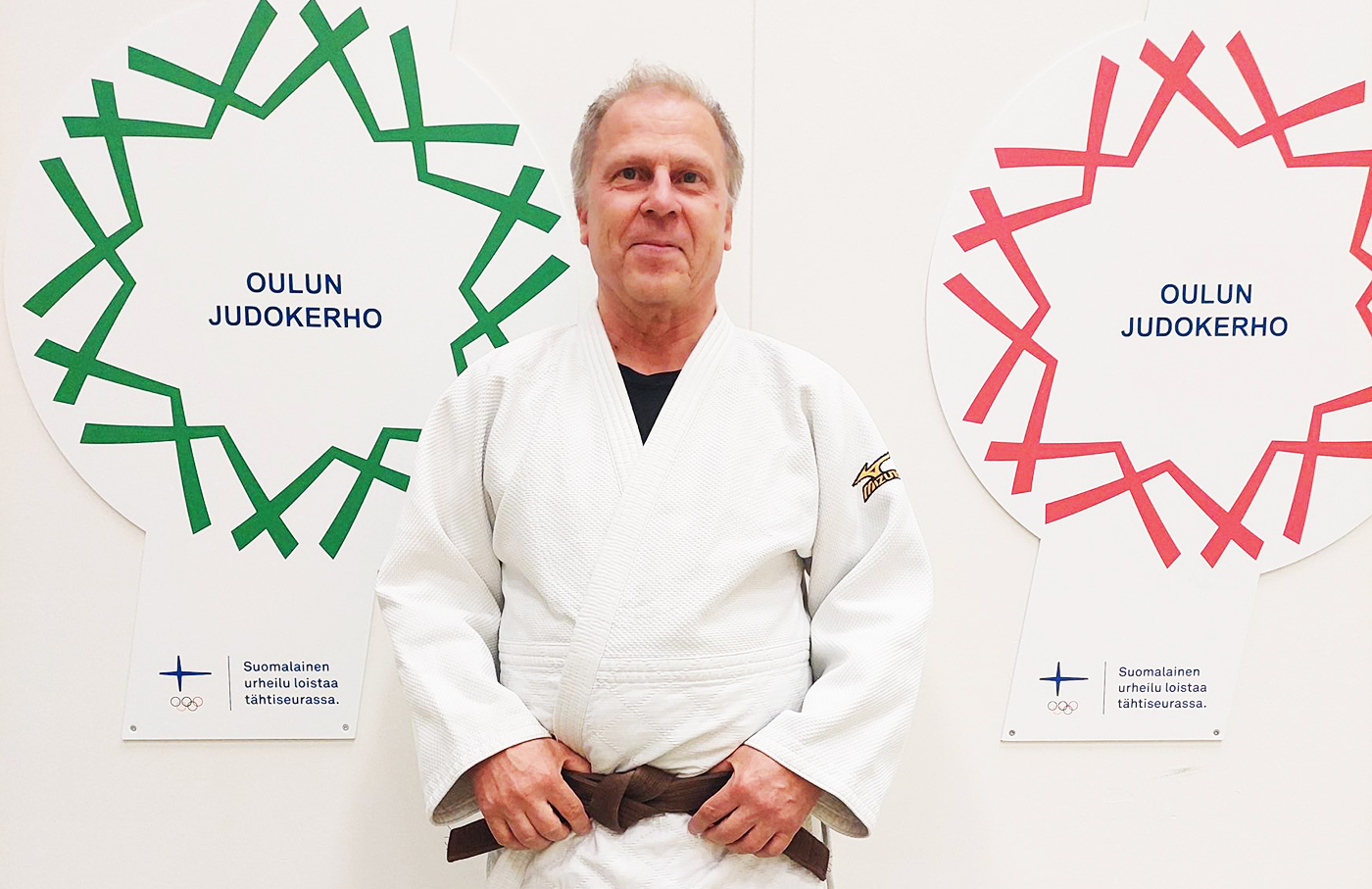 Arto Kallio poseeraamassa kameralle ruskea vyö vyötäröllään. Taustalla Oulun Judokerho-taulut. 